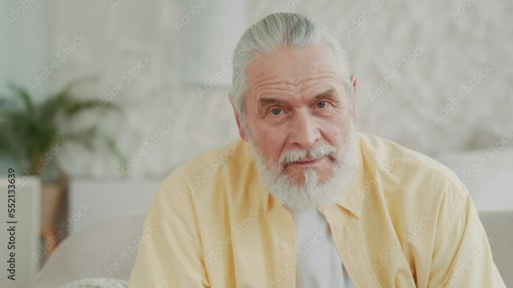 Close up portrait of older senior adult man looking at the camera sitting on sofa at home. Smiling. Relaxing. Indoor. Modern apartment. Close up portrait of older senior adult man looking at the camera sitting on sofa at home. Smiling. Relaxing. Indoor. Modern apartment.