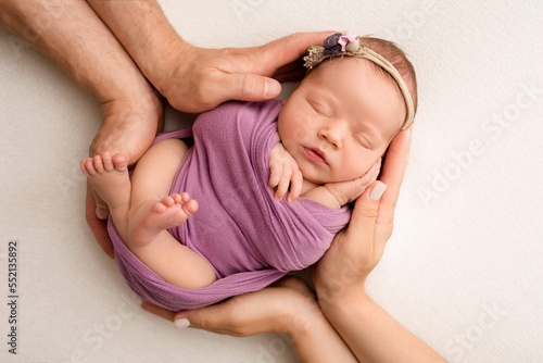Sleeping newborn girl in the first days of life on a white background. A newborn baby in a purple lilac winding and a headband. Hand, palms of father and mother, parents hold the child.