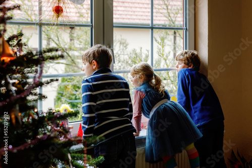 Three cute children sitting by window on Christmas eve. Two school kid boys and toddler girl, siblings looking outdoor and dreaming. Family happiness on traditional holiday