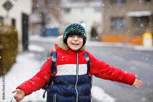 Little school kid boy of elementary class walking to school during snowfall. Happy child having fun and playing with first snow. Student with eye glasses backpack in colorful winter clothes.