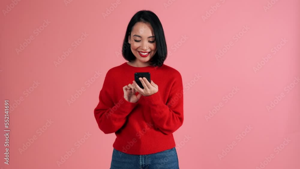 Smiling Asian woman wearing red sweater typing on mobile in the pink studio Smiling Asian woman wearing red sweater typing on mobile in the pink studio