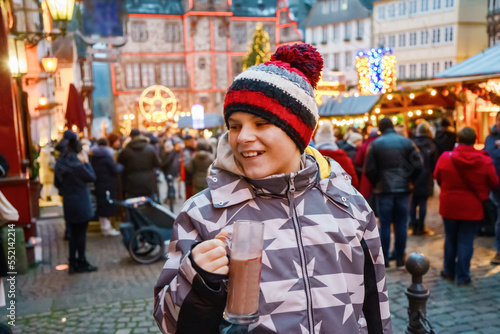 Little cute kid boy drinking hot children punch or chocolate on German Christmas market. Happy child on traditional family market in Germany, Laughing boy in colorful winter clothes
