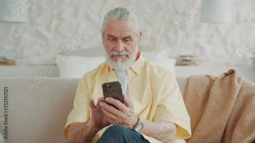 Happy attractive senior man uses a smartphone, browses and reads share text messages. Sitting on sofa at home looks at screen, swipes. Portrait