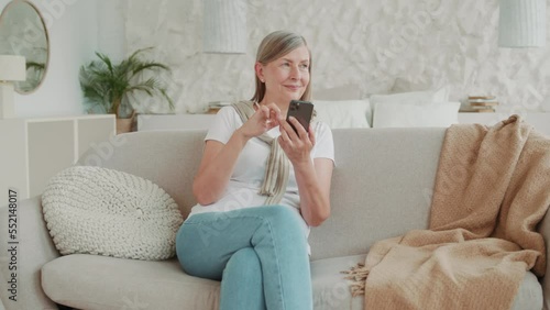 Beautiful smiling elder middle age woman using smartphone sitting on sofa at home in modern apartment. Feel happy. Communication. Mobile. Internet. Cellphone message