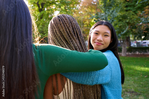 Affective photo of three female friends embracing outdoors at a public park, while one of them turns to look at camera smiling. Mixed race group of girls enjoying the sun at natural park. photo