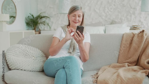 Elder middle age woman using smartphone sitting on sofa smiling at home in sunlight modern apartment. Communication. Mobile. Internet. Cellphone message
