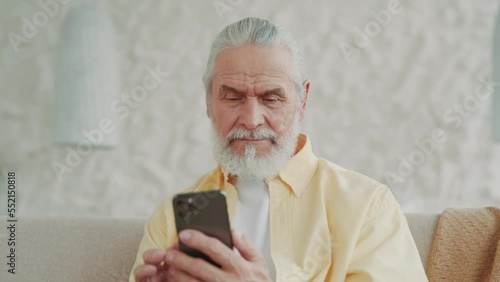 Close up senior man uses a smartphone, browses and reads share text messages. Sitting on sofa at home looks at screen, swipes. Portrait