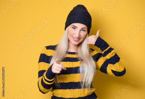 Call me. Woman gesturing at speaking on phone while looking to camera, model wearing woolen cap and sweater, isolated on yellow background. Woman on phone