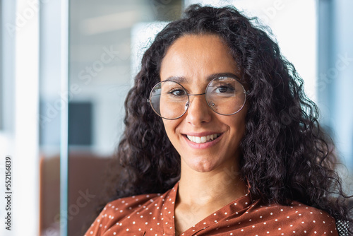 Fototapeta Naklejka Na Ścianę i Meble -  Close up photo portrait of beautiful Latin American woman with curly hair and glasses, businesswoman inside office building smiling and looking at camera.