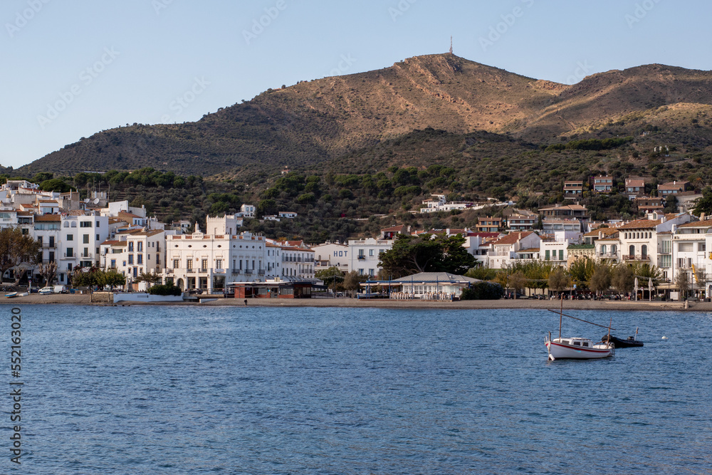 Barquita sobre el tranquilo mar Mediterráneo con las blancas casas del pueblo de Cadaqués y las verdes montañas de fondo.