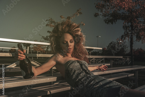 Vintage portrait of a tipsy beautiful young woman wearing an old school denim dress and long teased hair, celebrating alone with champagne in the bleachers of a soccer field