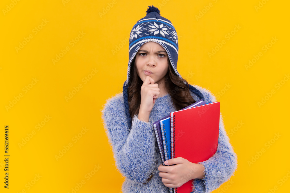 Thinking pensive clever teenager girl. School children in winter hat and sweater with books on isolated yellow studio background. Children learning and education. Thinking pensive clever teenager girl. School children in winter hat and sweater with books on isolated yellow studio background. Children learning and education.