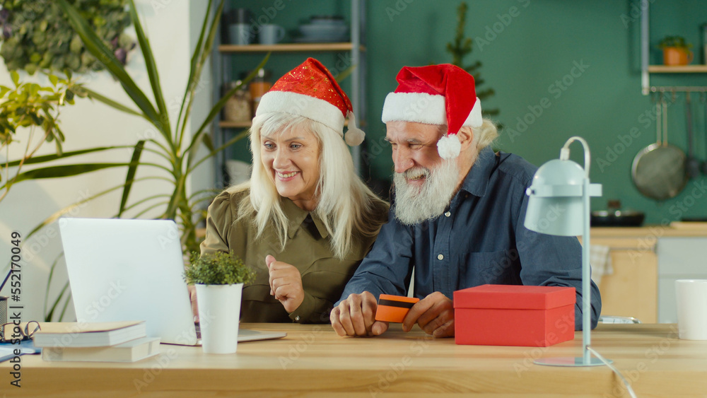 Excited happy elderly couple in Santa Claus hats are choosing Christmas gifts and shopping online using laptop and credit card at home. Holiday shopping and online shopping concept. Excited happy elderly couple in Santa Claus hats are choosing Christmas gifts and shopping online using laptop and credit card at home. Holiday shopping and online shopping concept.