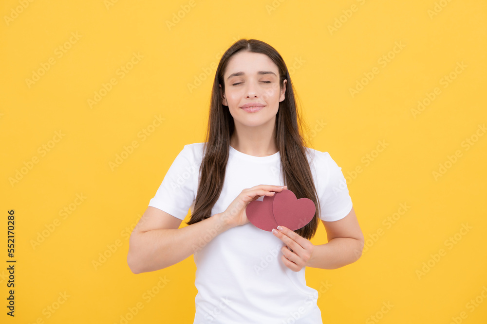 Romantic lovely girl. Valentines Day. Beautiful young woman with heart in her hands. Attractive woman with smile having red hearts in hands, closing eyes with paper heart over yellow background. Romantic lovely girl. Valentines Day. Beautiful young woman with heart in her hands. Attractive woman with smile having red hearts in hands, closing eyes with paper heart over yellow background.