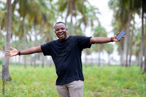 Cheerful smiling black african young man in village in Africa pointing finger at blank screen smartphone, for smart mobile application presentation projects.