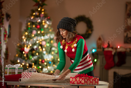 Serious young woman wrapping Christmas gifts in front of a Christmas Tree in her living room. photo