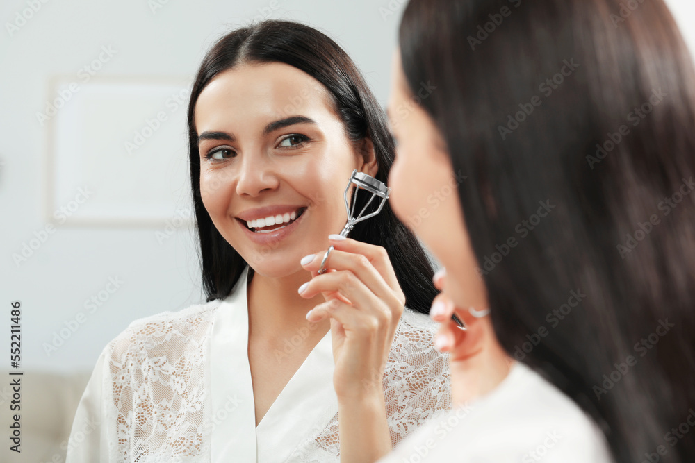 Beautiful young woman with eyelash curler near mirror indoors Beautiful young woman with eyelash curler near mirror indoors