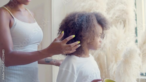 Cute Black girl standing drinking tea while mom making her hair, stroking daughters head gently. Thoughtful child touching soft toy and looking in window. Static shot. Childhood, parenthood concept.