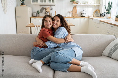Carefree happy family of millennial woman and teenage girl hugging sits on big sofa in living room. Positive cheerful daughter hugging mother after long separation or arrival from school camp