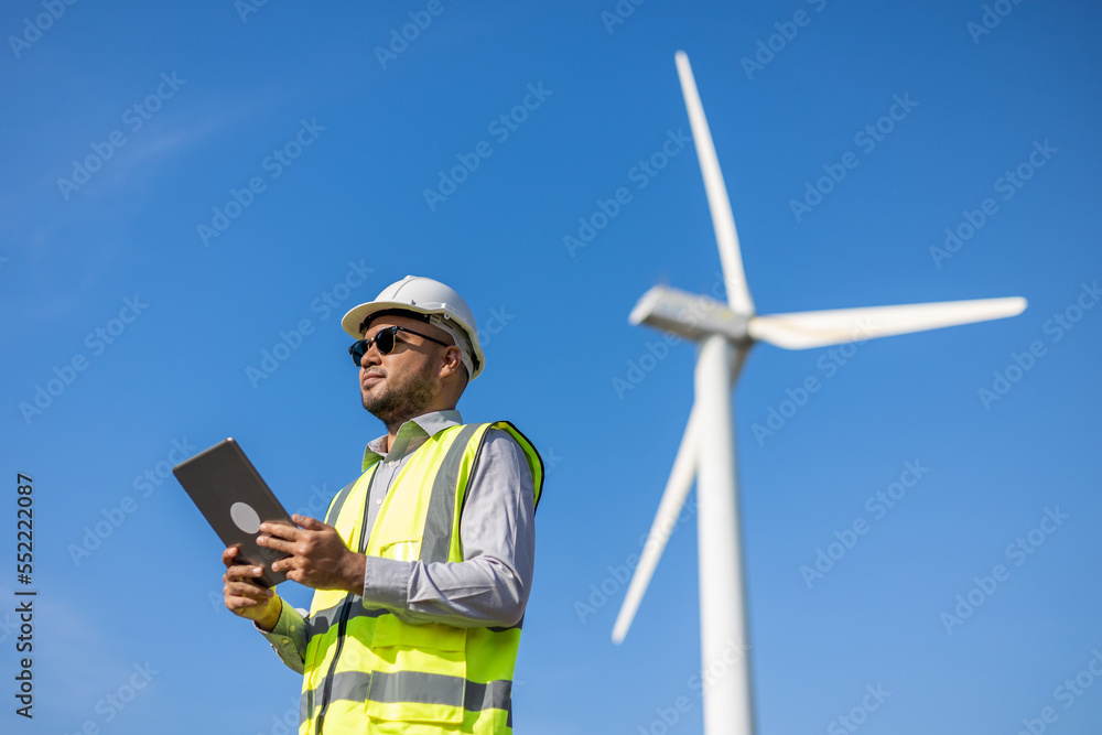 Engineer India man working with tablet at windmill farm Generating electricity clean energy. Wind turbine farm generator by alternative green energy. Asian engineer checking control electric power Engineer India man working with tablet at windmill farm Generating electricity clean energy. Wind turbine farm generator by alternative green energy. Asian engineer checking control electric power