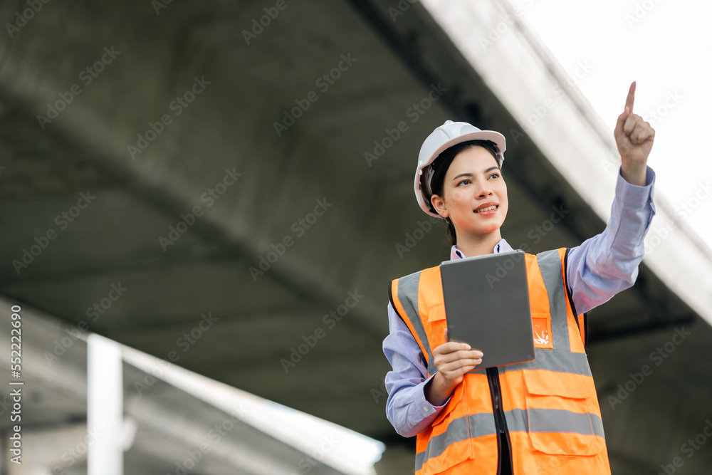 Asian engineer worker woman or architect looking construction with tablet wear white safety helmet in construction site. Standing at highway concrete road site. Progress planning of highway bridge. Asian engineer worker woman or architect looking construction with tablet wear white safety helmet in construction site. Standing at highway concrete road site. Progress planning of highway bridge.