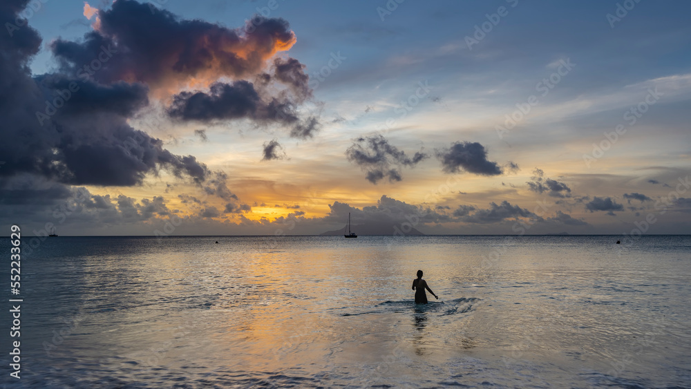 Silhouette of a girl in the ocean at sunset. There are picturesque clouds in the sky, highlighted in orange and pink. Reflection on the water. The silhouette of the island on the horizon. Seychelles.