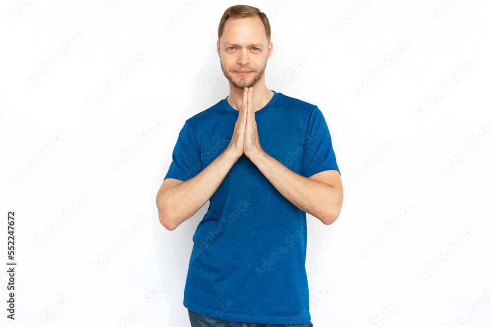 Peaceful man with praying hands looking at camera. Cheerful man in T-shirt looking at camera while standing on white background, praying, making wish or meditating.Harmony, hope, concentration concept Peaceful man with praying hands looking at camera. Cheerful man in T-shirt looking at camera while standing on white background, praying, making wish or meditating.Harmony, hope, concentration concept