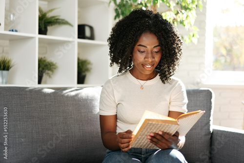 African woman reading book at home sitting on sofa