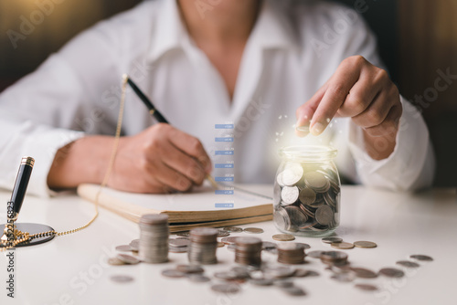 Businessman holding coins putting in jar and taking notes , Stacking coins, graph on table. plan revenue and expenses, target success growth, finance profit planning 2023 concept.