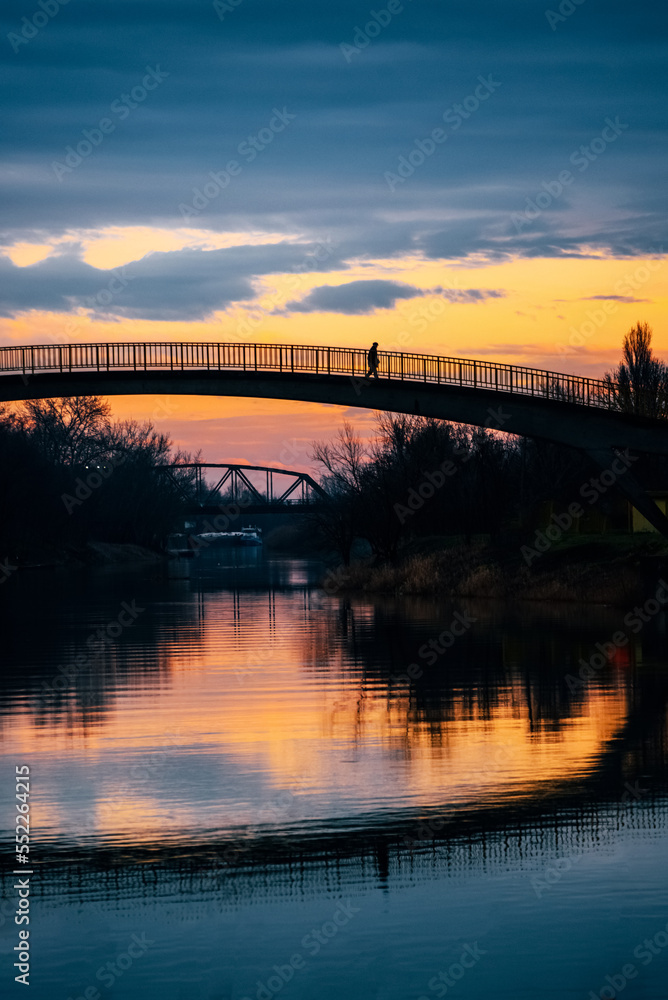 Silhouette of a man walking across small bridge over a peaceful river at sunset. Serene scenery in Zrenjanin city in Serbia on the river Begej