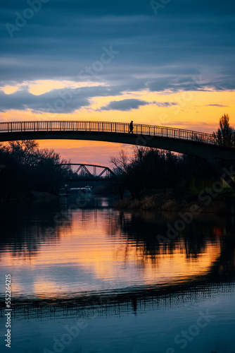 Fototapeta Naklejka Na Ścianę i Meble -  Silhouette of a man walking across small bridge over a peaceful river at sunset. Serene scenery in Zrenjanin city in Serbia on the river Begej