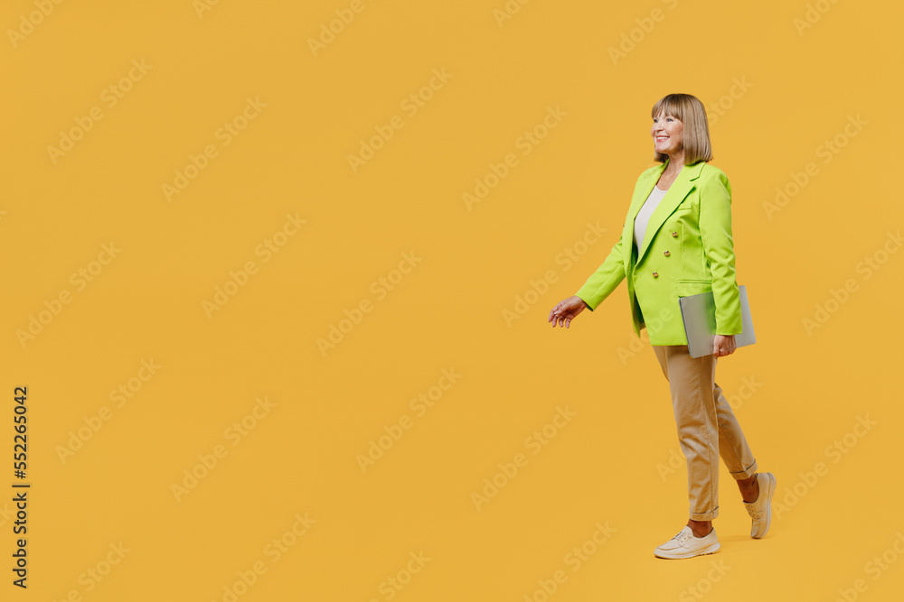Fototapeta premium SIde view full body smiling elderly IT woman 50s years old wears green jacket white t-shirt hold closed laptop pc computer isolated on plain yellow background studio portrait People lifestyle concept.