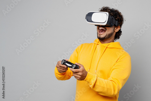 Young Indian man 20s he wearing casual yellow hoody hold in hand play pc game with joystick console watching in vr headset pc gadget isolated on plain grey background studio People lifestyle portrait