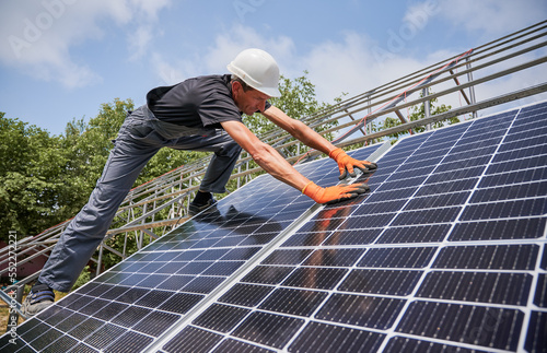 Man in safety helmet and work overalls building photovoltaic solar panel system outdoors. Male worker in orange gloves placing solar modules on metal rails. Concept of alternative energy sources.