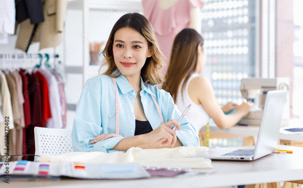 Obraz premium Millennial Asian young beautiful professional female designer dressmaker businesswoman measure fabric by measuring tape on working desk while colleague using sewing machine in tailor workshop studio