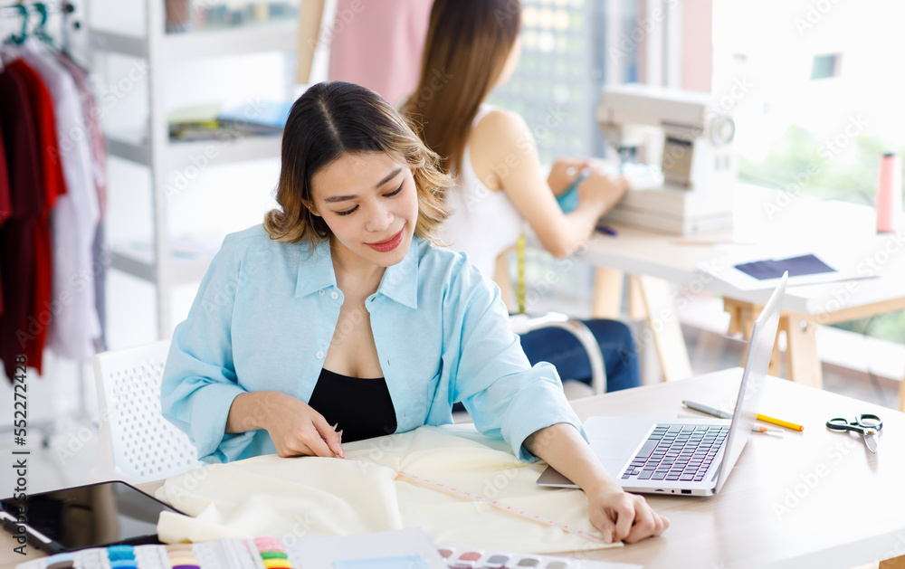 Millennial Asian young beautiful professional female designer dressmaker businesswoman measure fabric by measuring tape on working desk while colleague using sewing machine in tailor workshop studio Millennial Asian young beautiful professional female designer dressmaker businesswoman measure fabric by measuring tape on working desk while colleague using sewing machine in tailor workshop studio