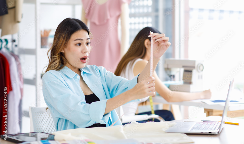 Asian young professional excited shocked female designer dressmaker businesswoman make wrong mistake with measuring tape on working desk when colleague using sewing machine in tailor workshop studio Asian young professional excited shocked female designer dressmaker businesswoman make wrong mistake with measuring tape on working desk when colleague using sewing machine in tailor workshop studio