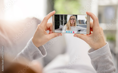Mature woman lying on comfy couch and holding smartphone with video call, pleasant conversation with friend. Distancing video chat
