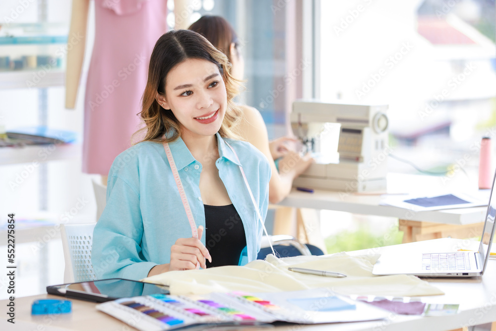 Millennial Asian young beautiful professional female designer dressmaker businesswoman measure fabric by measuring tape on working desk while colleague using sewing machine in tailor workshop studio