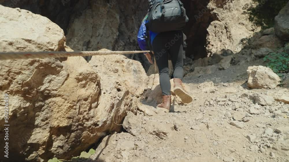 Back view of woman hiking and backpack in blue jacket wearing boots was hands to hold the rope up the mountain. Back view of woman hiking and backpack in blue jacket wearing boots was hands to hold the rope up the mountain.