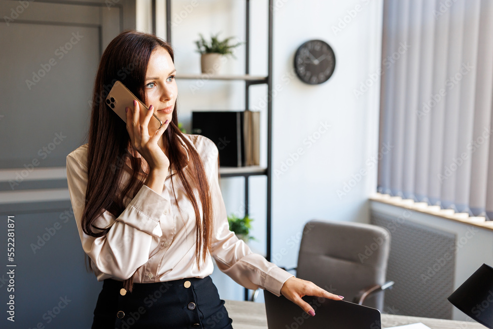 Business talk. Cheerful young beautiful woman talking on mobile phone and looking to the window while leaning on the table at her working place Business talk. Cheerful young beautiful woman talking on mobile phone and looking to the window while leaning on the table at her working place
