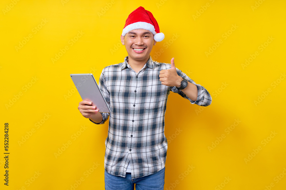 Young handsome smiling Asian man wearing Christmas hat holding tablet showing thumb up sign on yellow background. Celebration Christmas and Happy New Year 2023 holiday concept