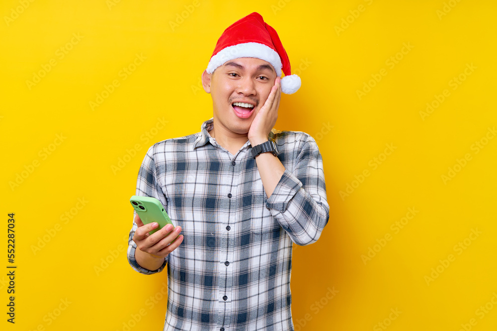 Excited young Asian man in plaid shirt wearing Christmas hat holding mobile phone and putting hand on his cheek looking camera on yellow background. Christmas and Happy New Year 2023 holiday concept Excited young Asian man in plaid shirt wearing Christmas hat holding mobile phone and putting hand on his cheek looking camera on yellow background. Christmas and Happy New Year 2023 holiday concept