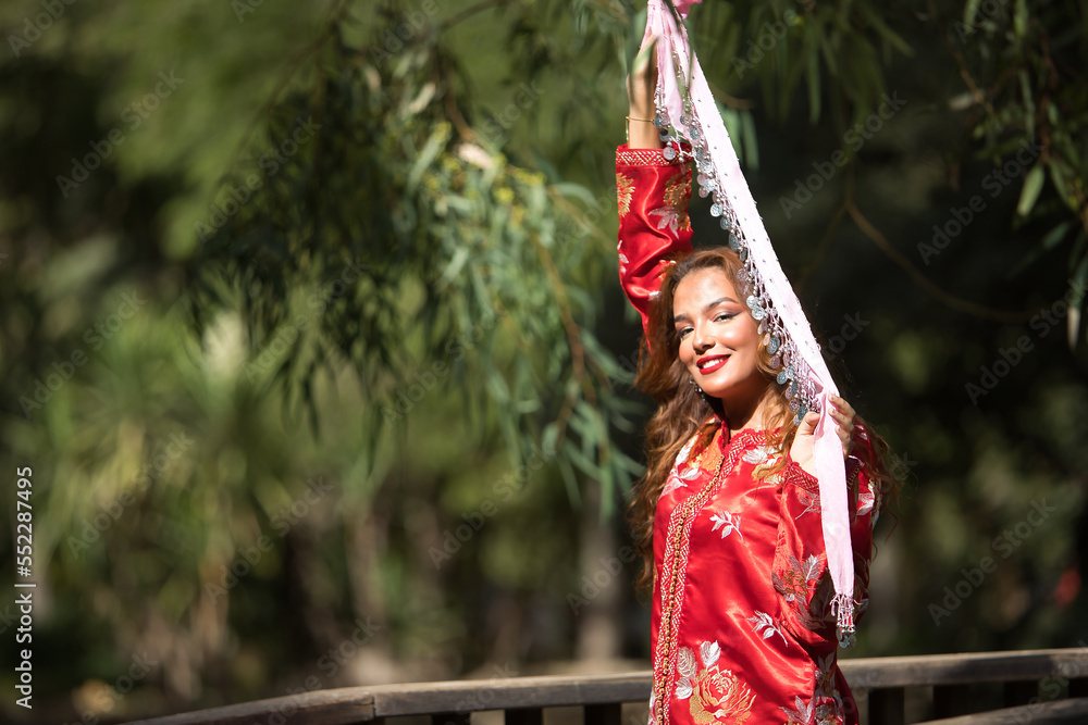 Obraz premium Beautiful young woman in a typical Moroccan red suit, embroidered with gold and silver threads, holding a scarf with coins in her hands, on a wooden bridge. Concept beauty, ethnicity, Marrakech, Arab