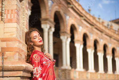 Young and beautiful woman in a typical Moroccan red costume, with embroidery with gold and silver threads, leaning on a brick column in the Spanish square. Concept ethnicity, typical costumes, Arabic