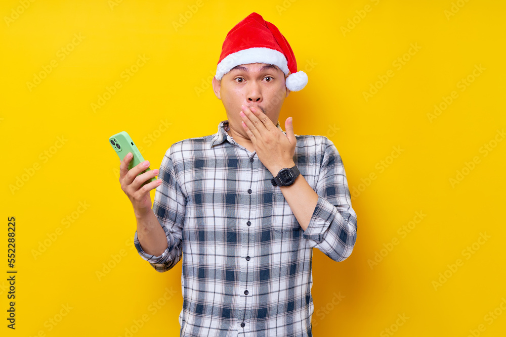 surprised young Asian man in plaid shirt wearing Christmas hat holding smartphone and posing cover mouth with hand on yellow background. Christmas and Happy New Year 2023 holiday concept surprised young Asian man in plaid shirt wearing Christmas hat holding smartphone and posing cover mouth with hand on yellow background. Christmas and Happy New Year 2023 holiday concept