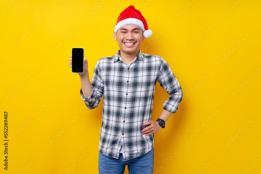 Smiling young Asian man in plaid shirt wearing Christmas hat holding mobile phone with blank screen workspace area on yellow background. Christmas and Happy New Year 2023 holiday concept