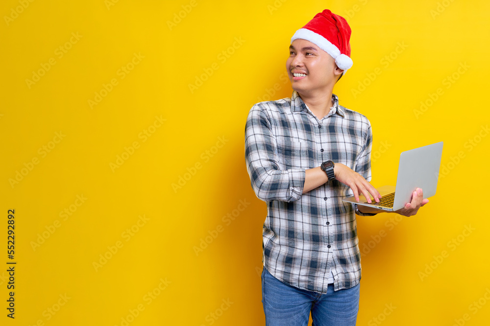 Smiling young Asian man in plaid shirt wearing Christmas hat using laptop pc computer on work and standing looking aside on yellow background. Christmas and Happy New Year 2023 holiday concept Smiling young Asian man in plaid shirt wearing Christmas hat using laptop pc computer on work and standing looking aside on yellow background. Christmas and Happy New Year 2023 holiday concept