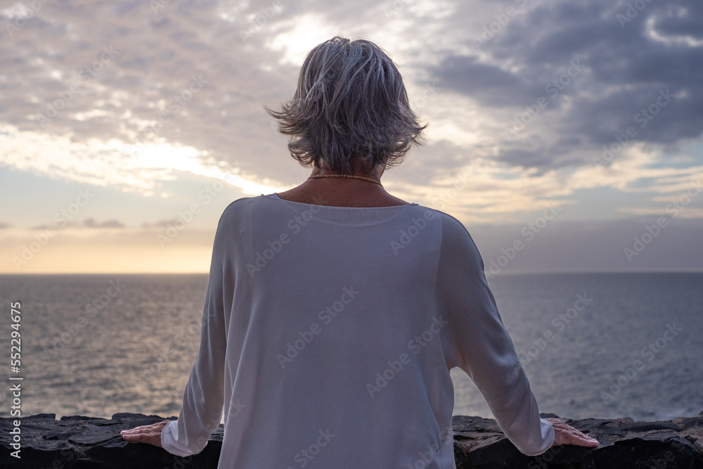 Fototapeta premium Rear view of senior woman standing in front of the sea at sunset looking at the horizon over the water. Peace, solitude, relaxation, meditation