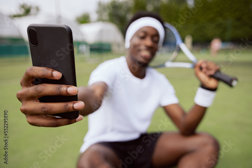 Close-up of hand of African-looking man holding phone in black case. The guy is wearing white t-shirt and headband, smiling and taking a selfie. He holds tennis racket in hand.